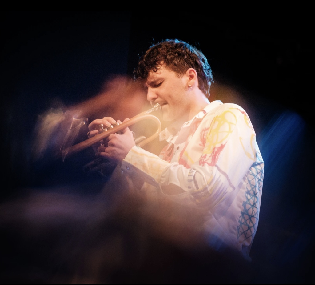 Der junge Trompeter Lorenz Widauer vor schwarzem Hintergrund. Er trägt kurze Haare und ein weißes Hemd mit bunten Stickereien. Das Foto ist durch den Effekt der Langzeitbelichtung stilisiert.