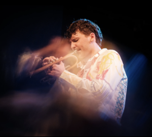 Der junge Trompeter Lorenz Widauer vor schwarzem Hintergrund. Er trägt kurze Haare und ein weißes Hemd mit bunten Stickereien. Das Foto ist durch den Effekt der Langzeitbelichtung stilisiert.