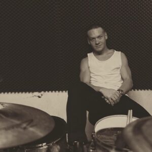 Der junge Schlagzeuger Paul Widauer sitzt locker mit dem Rücken an eine Wand gelehnt in seinem Schlagzeugsessel. Er trägt ein weißes Tanktop. Vor ihm sieht man Teile seines Schlagzeugs.